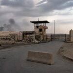 Base militare nel deserto con atmosfera di tensione e lieve fumo sullo sfondo.