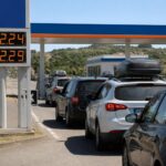Auto in coda a un distributore in autostrada durante l'esodo estivo, con prezzi dei carburanti sopra i 2 euro al litro.