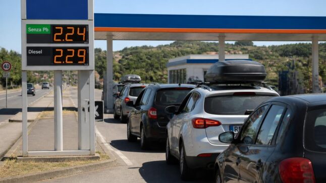 Auto in coda a un distributore in autostrada durante l'esodo estivo, con prezzi dei carburanti sopra i 2 euro al litro.